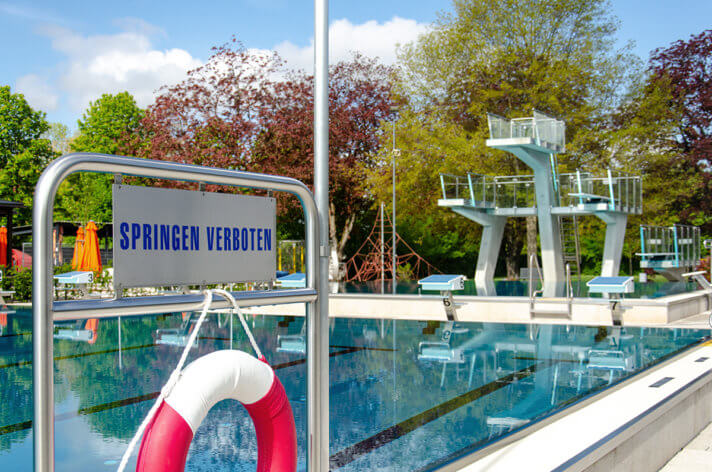Foto: Im Vordergrund steht das Schild: Springen verboten. Im Hintergrund ist ein Teil des Schwimmerbeckens und der 5-Meter Sprungturm zu sehen.