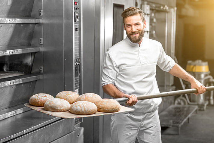 Bäcker schiebt ein Brot in den Ofen und schaut dabei freundlich in die Kamera..
